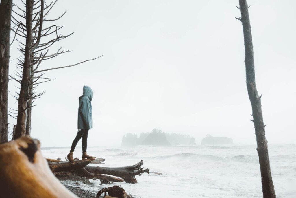 Full body of unrecognizable female in hood standing on log near leafless trees on wet shore near stormy sea in foggy weather