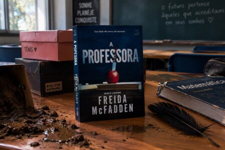 Capa do livro "A professora", de Freida McFadden com um fundo de sala de aula. Elementos presentes: caixas de sapato, lama e livros